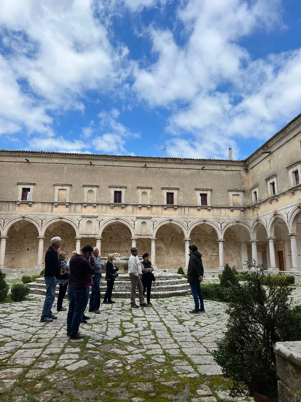Un gruppo in visita ascolta una persona che racconta, in un cortile storico circondato da arcate in pietra.